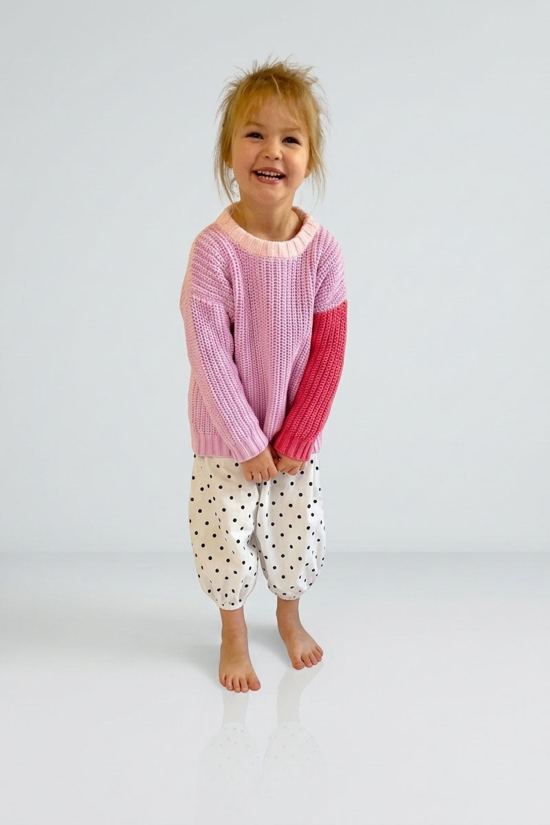 Child wearing a pink and red sweater with white polka dot pants on a light grey background