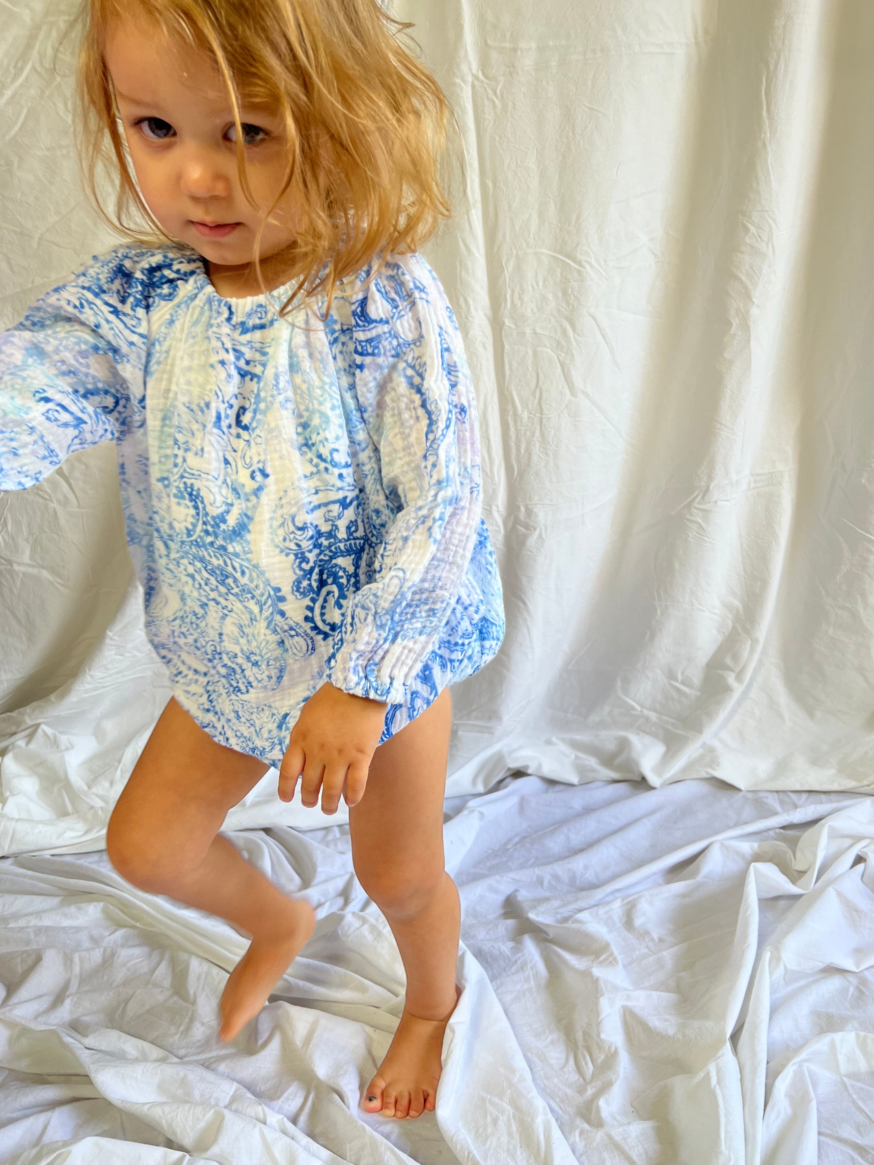 Young child stood in front of a white background  wearing a Long Sleeve Romper in Blue Paisley colour
