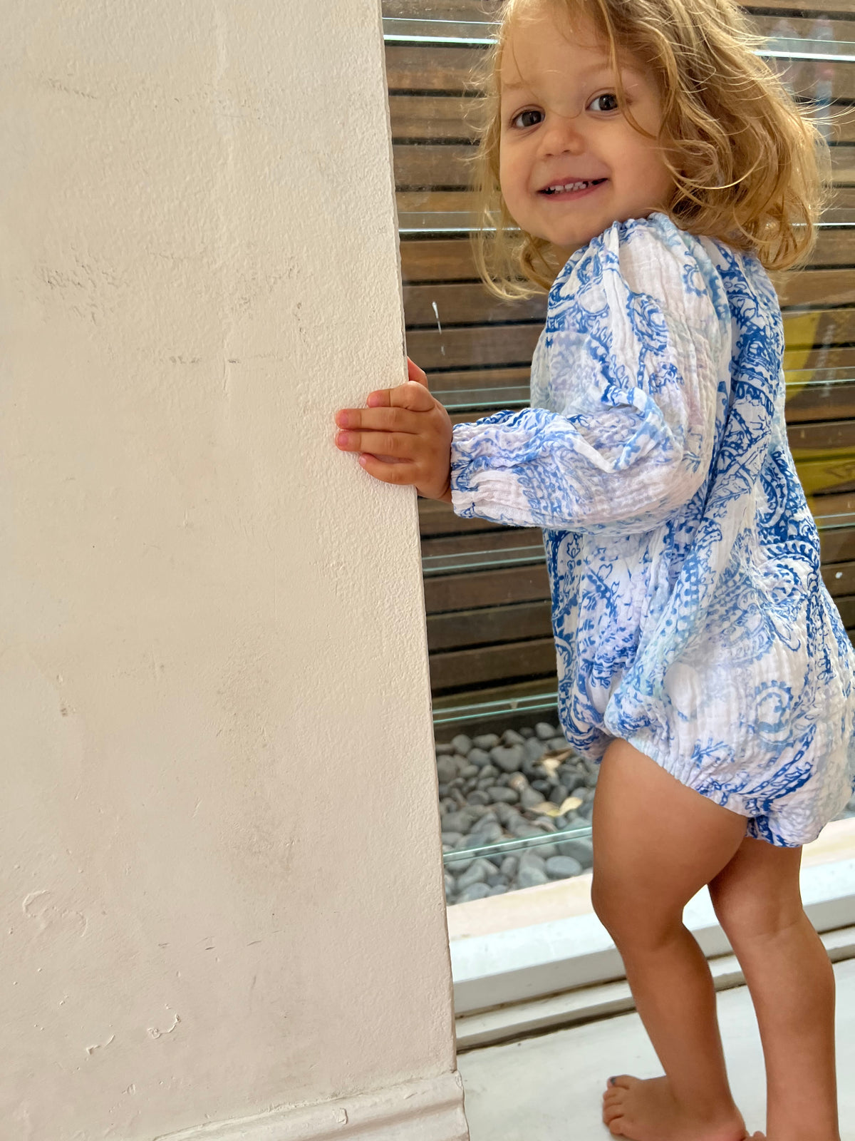 Young child stood in front of a window smiling wearing a Long Sleeve Romper in Blue Paisley colour