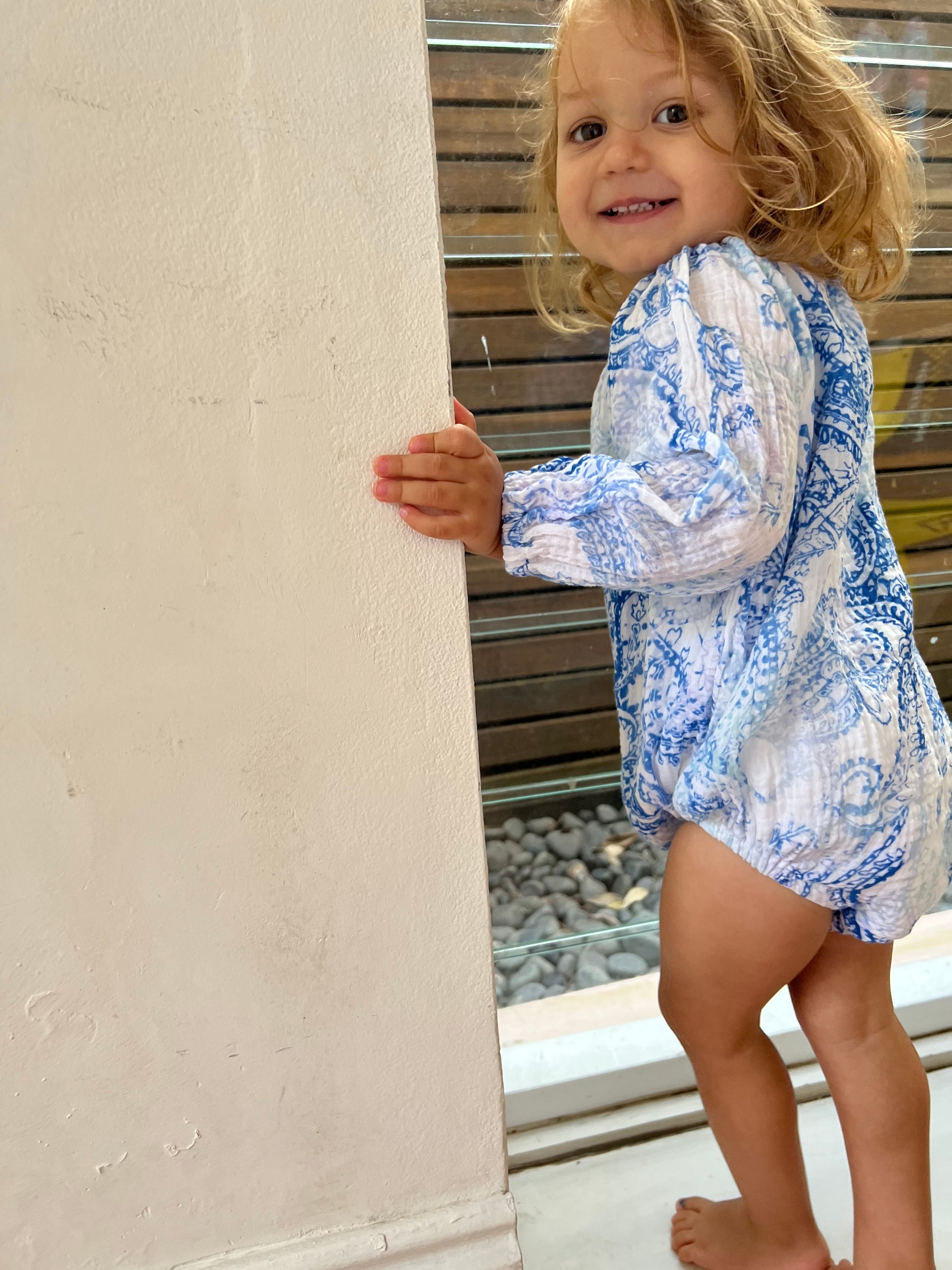 Young child stood in front of a window smiling wearing a Long Sleeve Romper in Blue Paisley colour