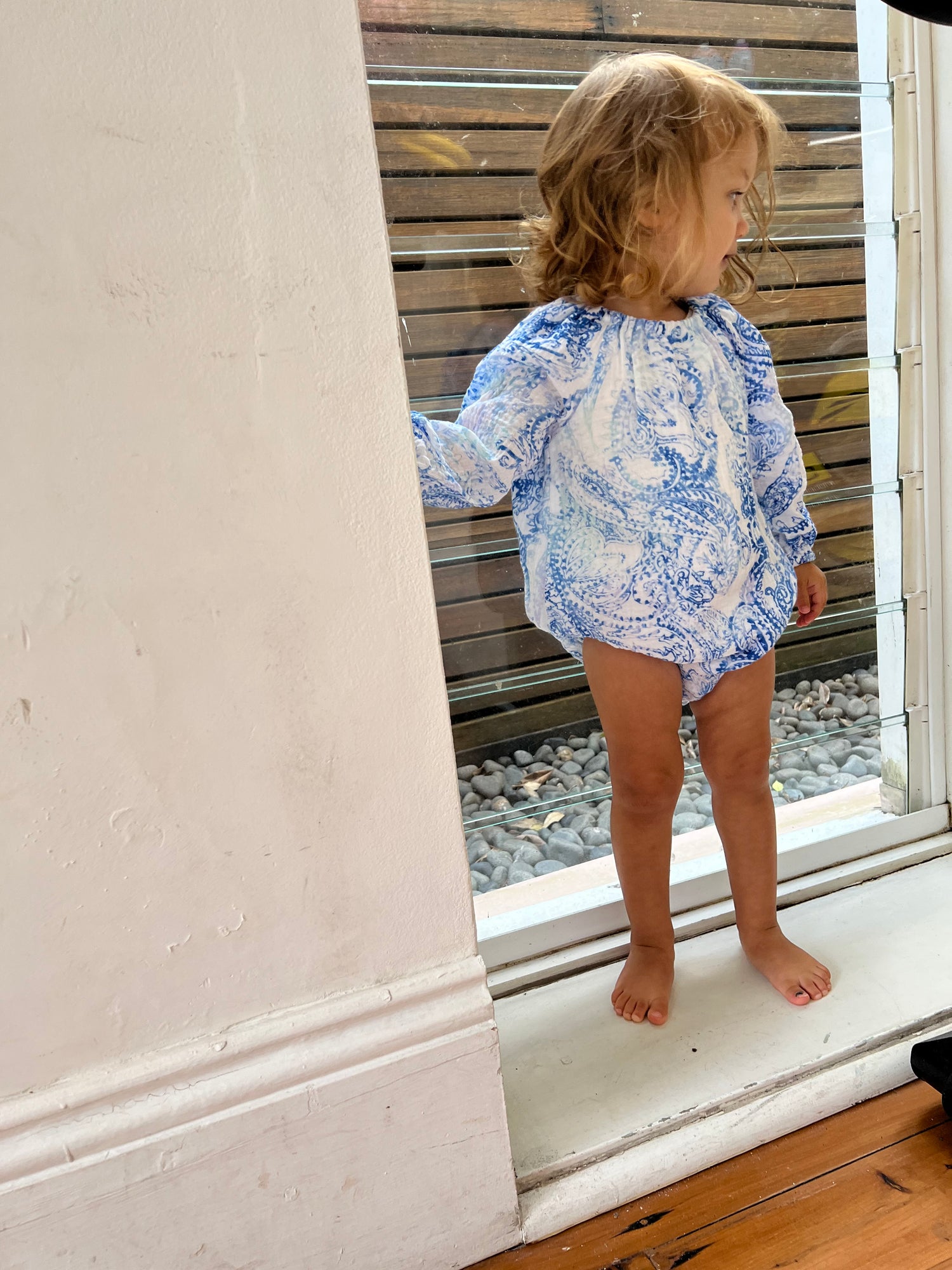 Young child stood by a window wearing a Long Sleeve Romper in Blue Paisley colour