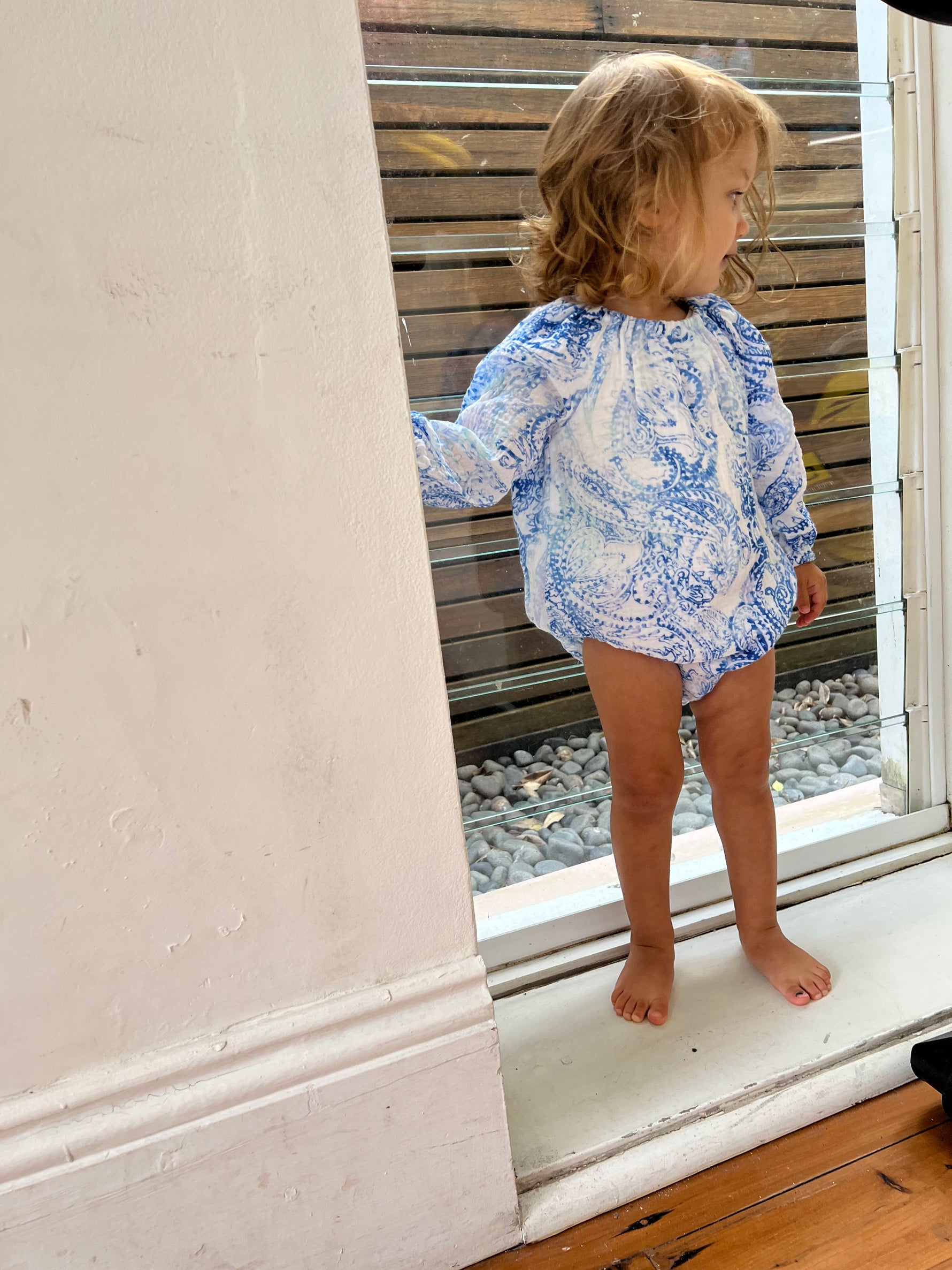 Young child stood by a window wearing a Long Sleeve Romper in Blue Paisley colour