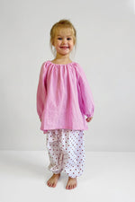 Child wearing a pink long sleeve top and white pants with polka dots on a plain background