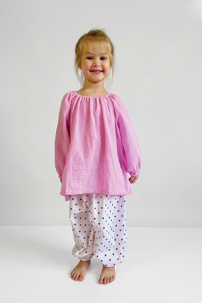Child wearing a pink long sleeve top and white pants with polka dots on a plain background