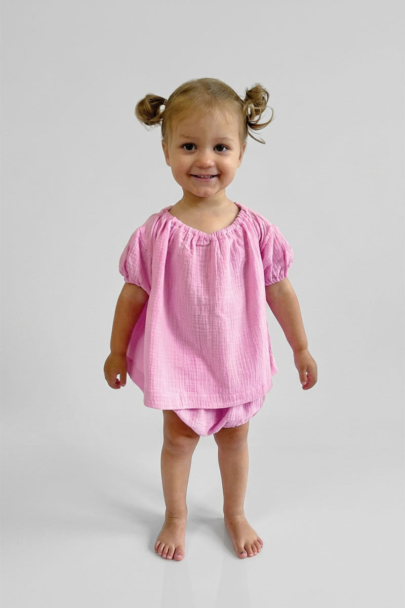 Young girl wearing a pink short sleeve puff top and matching pink bloomers on a plain background