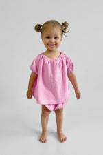 Young girl wearing a pink short sleeve puff top and matching pink bloomers on a plain background