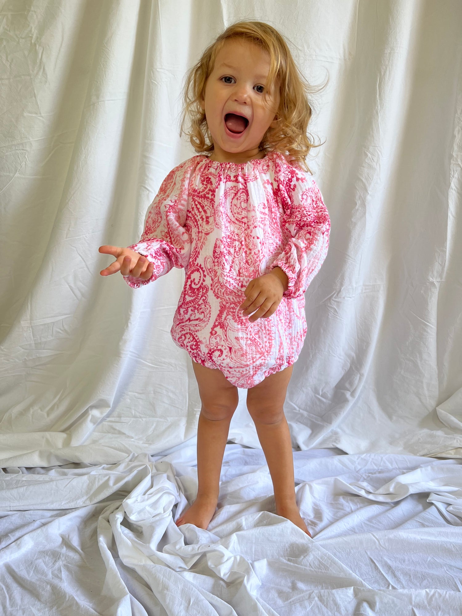 Young child smiling while wearing a Long Sleeve Romper in Paisley Red colour